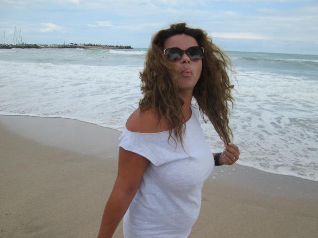 Mujer con cabello castaño ondulado, vistiendo una blusa blanca con un hombro descubierto, gafas de sol y un reloj de pulsera, de pie en una playa arenosa con olas suaves, con los labios fruncidos..