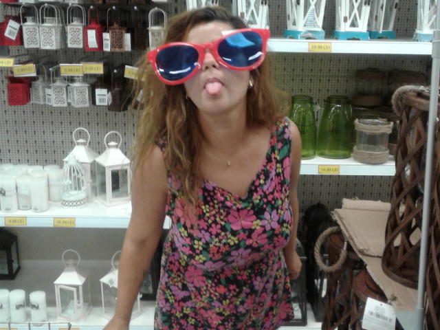 FacialEjac with wavy brown hair, pink floral dress, red oversized sunglasses, and tongue out, standing in a store, holding her hips, showing a cute, playful expression.