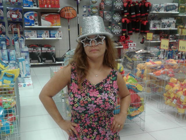 Mulher com cabelo castanho, usando um chapéu de estrela prateada e um vestido floral, em pé em uma loja de brinquedos, com as mãos nos quadris, mostrando os seios..