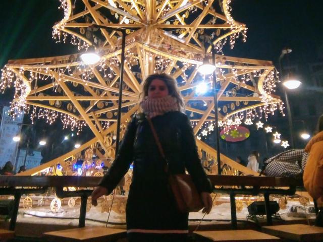FacialEjac with dark coat, white scarf, and brown purse stands in front of a brightly lit, golden star-shaped Christmas tree. Her face is slightly blurred, with a serious expression.
