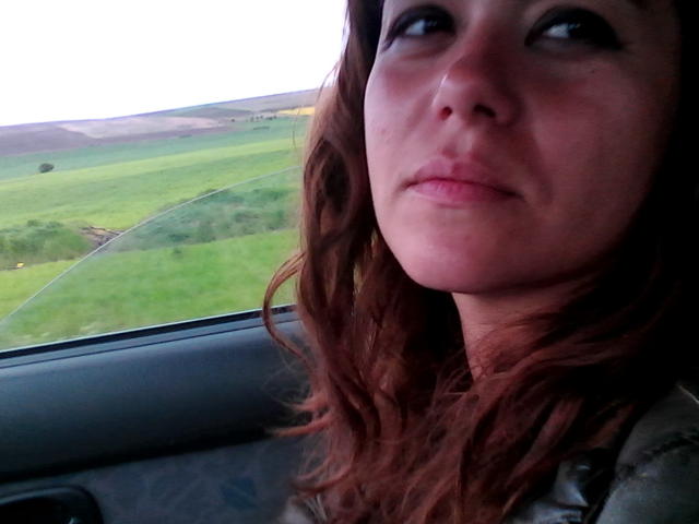 FacialEjac has wavy brown hair, a slightly flushed face, and a subtle smile. She's sitting in a car, looking out the window with green fields outside.