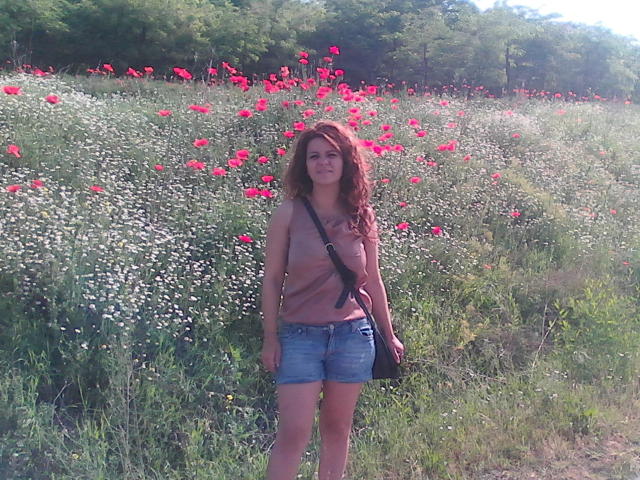 Een rode FacialEjac met golvend haar, kleine borsten en een zwarte schoudertas staat in een veld met roze en witte bloemen, gekleed in een roze top en denim shorts..