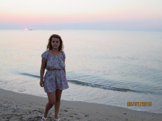 A curly-haired woman with a red headband stands on a sandy beach in a floral dress, white sneakers, and a serious expression. She has small tits and a cute ass.