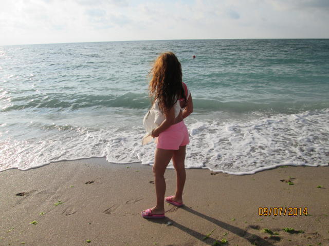 FacialEjac, una donna corpulenta con lunghi capelli ricci castani, è in piedi sulla spiaggia, indossa pantaloncini rosa e sandali rosa, guarda l'oceano e tiene in mano una borsa bianca..