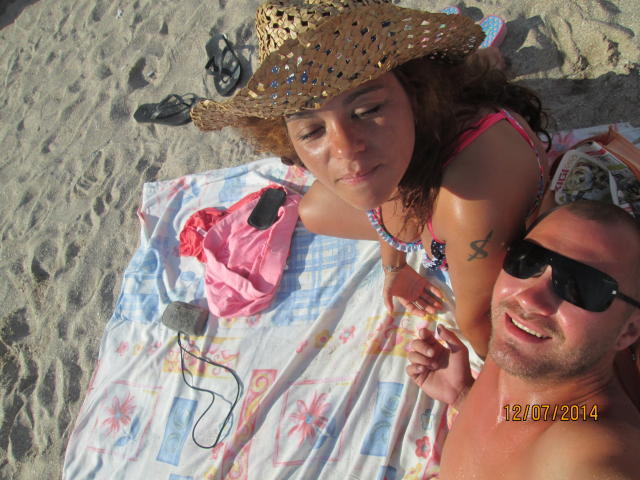 Van bovenaf genomen foto van een vrouw met bruin haar, bruine huid en kleine borsten in een roze bikini, met een strohoed, liggend op een strandlaken, samen met een man met een FacialEjac-shirt en zonnebril, die hand in hand liggen..