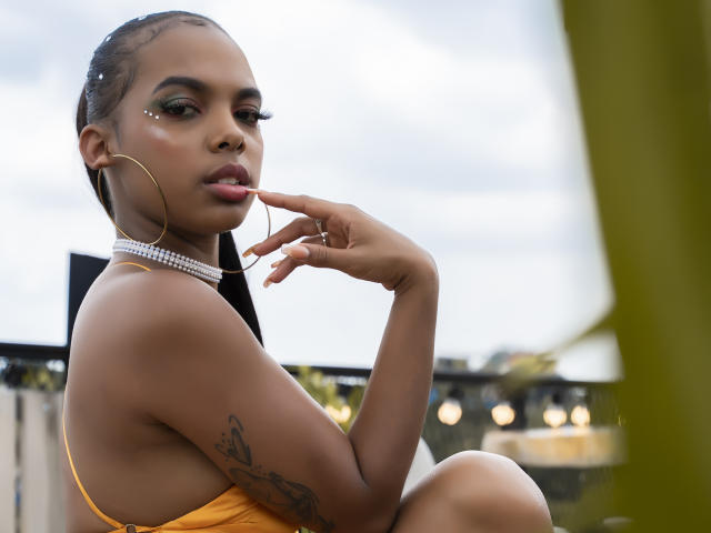 Leyah Hill, com pele escura, cabelo trançado, vestindo um vestido amarelo, brincos grandes em formato de argola e um colar de diamantes, chupando o dedo, mostrando uma tatuagem no braço superior..