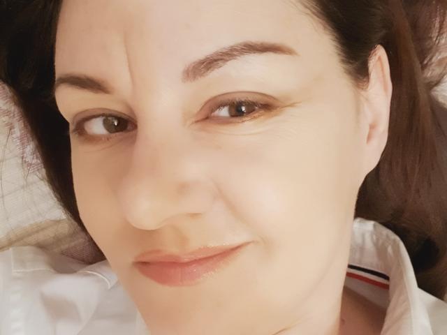 Il viso di Anna Fock, con un leggero sorriso, pelle chiara, occhi marroni e sopracciglia scure, indossa una camicia bianca con strisce rosse e nere, sdraiata..