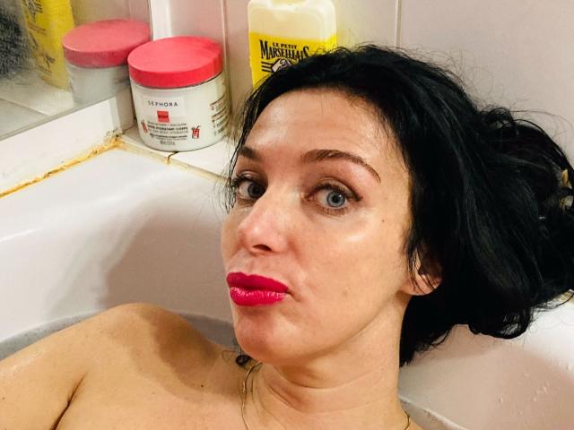 KatieFrenchie in a bathtub with wet black hair, red lipstick, and blue eyes, looking at the camera, with shampoo bottles and a yellow bottle in the background.