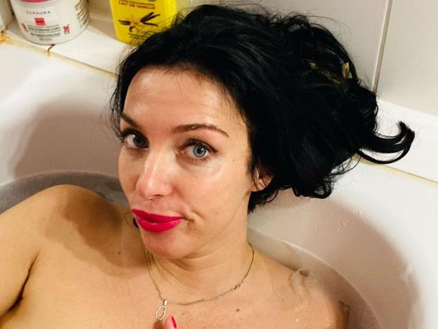 KatieFrenchie with dark messy hair, blue eyes, and bright pink lipstick, topless in a white bathtub, wearing a necklace with a pink charm, water up to her shoulders.