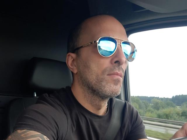 RichyLegend, kaal met een baard, draagt reflecterende zonnebril, een zwart T-shirt en een veiligheidsgordel, zit in een auto en kijkt vooruit met een lichte grijns..