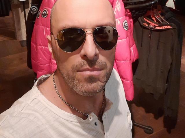 RichyLegend has a bald head, a beard, and wears aviator sunglasses with a white shirt and chain necklace. Pink puffy jacket is in the background.