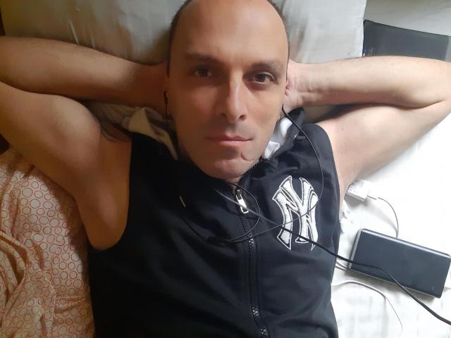 RichyLegend, calvo, indossa una felpa nera senza maniche con il logo bianco di NYC, è sdraiato su un letto con le braccia dietro la testa, ha le cuffie nelle orecchie e il telefono a destra..