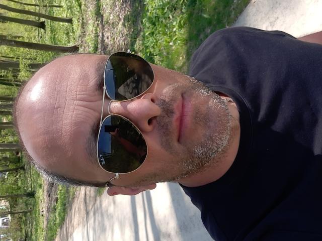 RichyLegend, mit kahlrasiertem Kopf, grauem Bart, trägt ein schwarzes T-Shirt und Aviator-Sonnenbrillen, steht in einem Wald, blickt in die Kamera, hält ein Telefon, das Sonnenlicht spiegelt sich in den Sonnenbrillen..