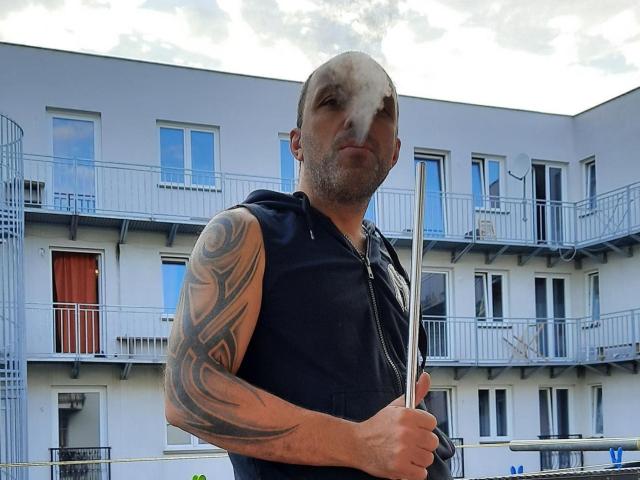 RichyLegend, con la testa calva, tatuaggi sul braccio destro, indossa una felpa nera senza maniche, tiene un bastone di legno e si trova di fronte a un edificio con balconi..