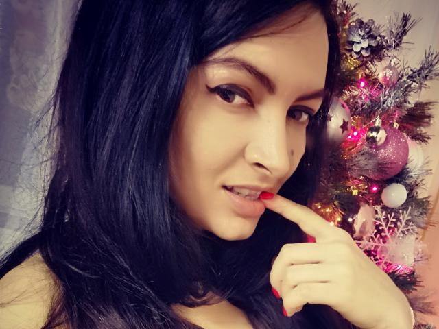 PassionX with black hair, red nails, and slightly parted lips, gently biting her finger. Christmas tree with pink and white ornaments in the background.