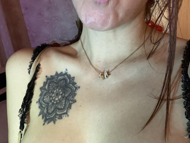 Misstyk's upper body with a black mandala tattoo on her left boob, wearing black lace straps and a gold butterfly necklace, puffy pink lips, wet brown hair.
