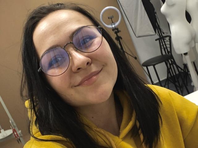 AlisaHottyLisa a la peau pâle, les cheveux noirs, et porte des lunettes rondes teintées de bleu avec un sweat à capuche jaune. Elle sourit légèrement, a un petit nez et pas de seins visibles..