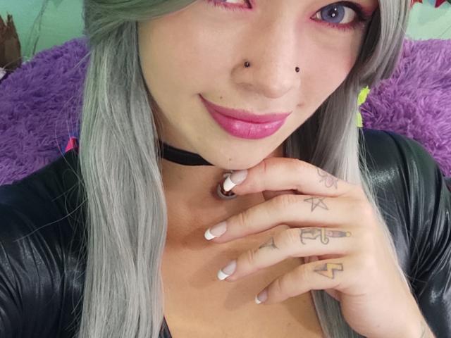 GoddessLufe has gray hair, pink eyelids, black nose ring, pink lipstick, tattoos on fingers, black choker, and is touching her chin with a black leather top.
