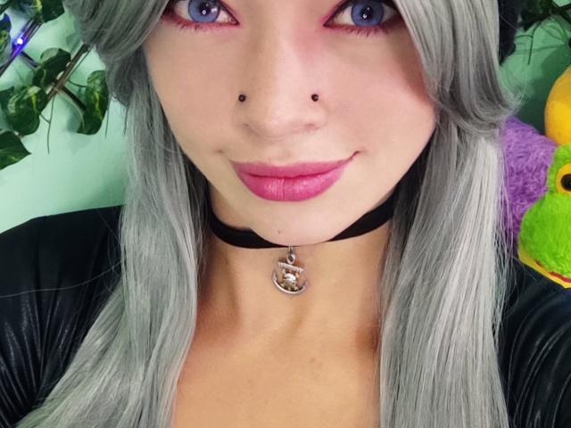 GoddessLufe con cabello plateado, ojos azules, lápiz labial rosa, collar negro con anillo, piercings en la nariz, blusa negra, sonriendo a la cámara, luciendo sexy y linda..
