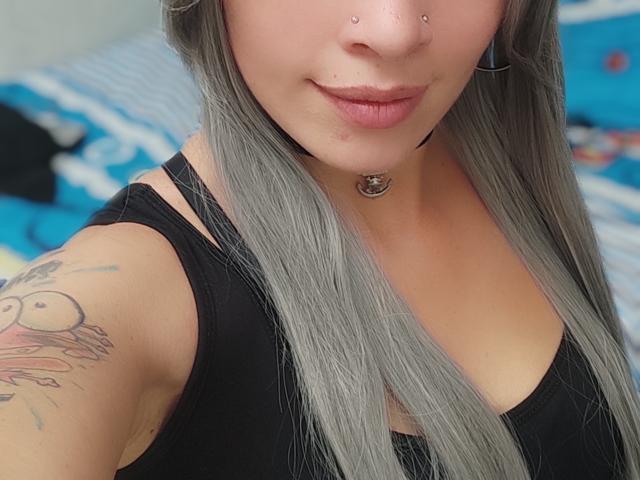 GoddessLufe, con cabello gris, un top de tirantes negro, piercings en la nariz, un aro en el labio, un collar y un tatuaje de una cara de dibujos animados en el brazo. Sus pechos son pequeños y está sonriendo..