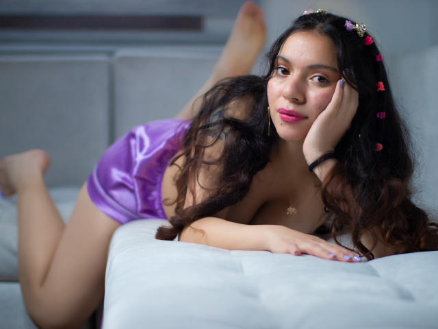 ElizabethBlizz, com cabelos longos e escuros, shorts de cetim roxo e grampos de cabelo com flores rosa, deitada em uma cama branca, com as pernas levantadas, a mão no rosto, sorrindo, mostrando os seios..