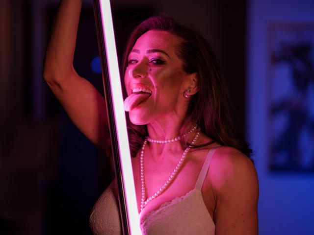 Joanna Braun, con la lengua afuera, sosteniendo un palo rosa neón, vistiendo un sujetador blanco de encaje y un collar de perlas, sonriendo, iluminación en tonos morado oscuro y rosa, cabello castaño, aretes..