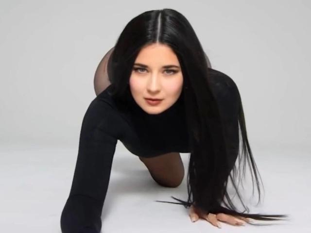 JudyLaVida is on all fours, wearing a black long-sleeve top, with long black hair, looking directly at the camera, her ass raised in the air.