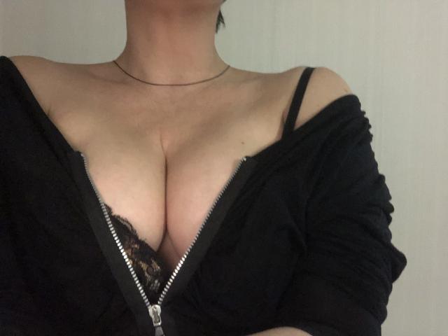LadyMiller's tits are showing through a black lace bra, zipped down in a black off-shoulder top, with a thin necklace around her neck.