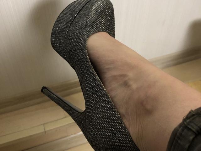 LadyMiller's foot in a sparkly black high heel, showing a visible blue bruise on her ankle. Her leg is slightly bent, with the heel pointed upwards.