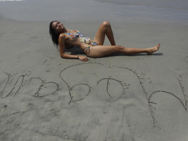 MeganGil deitada em uma praia arenosa, vestindo um biquíni colorido com estampas, pernas estendidas, braços atrás da cabeça, cabelo cacheado molhado, pele bronzeada, um pequeno coração e a palavra "AMOR" escritos na areia, sorrindo..