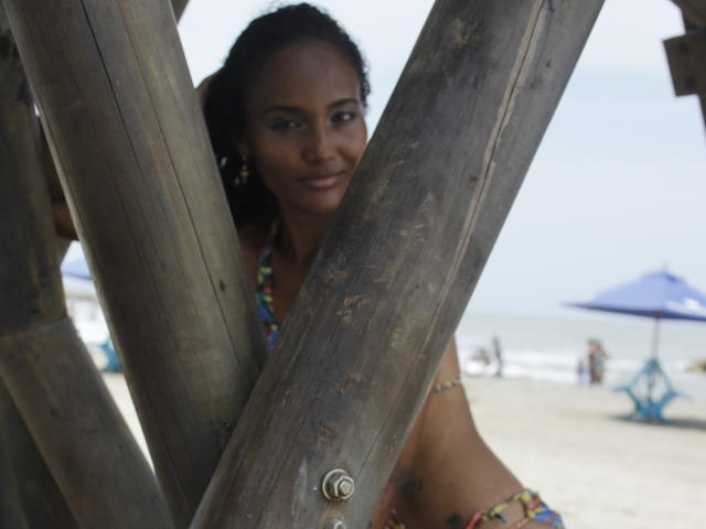 Megan Gil, con i capelli ricci e scuri, seno piccolo, indossa un bikini colorato e si trova dietro delle travi di legno, guardando la fotocamera con un sorriso appena accennato. Sullo sfondo, si vede la spiaggia..