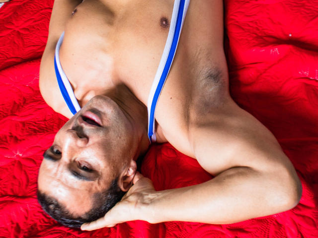 ToroHot, torse nu avec des bretelles rayées bleu et blanc, est allongé sur une courtepointe rouge, la tête penchée en arrière, les yeux à moitié ouverts, avec un regard sexy et un léger sourire, des bras musclés, des cheveux foncés..
