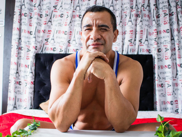 ToroHot, sin camisa, musculoso, piel bronceada, cabello negro corto, traje de baño azul, manos sobre la cama, sábanas rojas y blancas, hojas verdes sobre la cama, ligera sonrisa, confiado, sexy, atractivo..