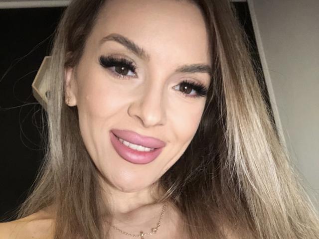 SophieDeee has long blonde hair, thick eyebrows, long eyelashes, pink lipstick, and a slight smile. Her face is centered, looking directly at the camera.