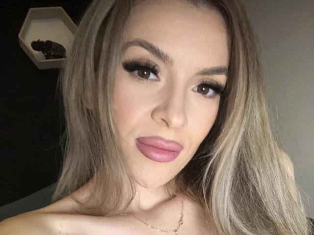 SophieDeee has long blonde hair, full pink lips, and thick black eyelashes. She's looking at the camera with a slight smirk, wearing a thin gold necklace.