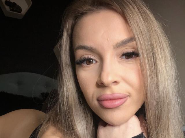 SophieDeee with pouty pink lips, long blonde hair, and thick black eyelashes, looking into the camera with a slight smile, her neck resting on her hand.