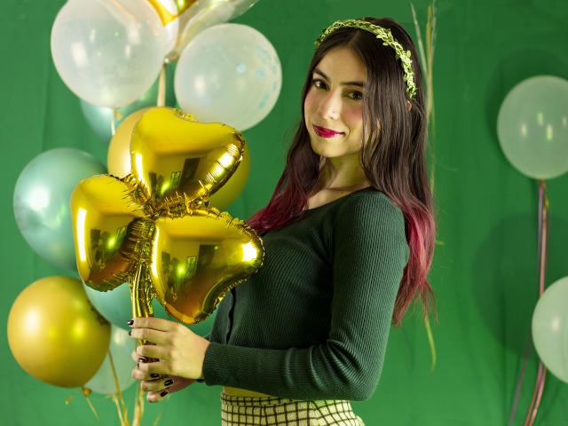 JuliaGarciax segurando balões dourados em formato de coração, vestindo um suéter verde, uma saia xadrez, uma tiara verde e batom rosa, com cabelos castanhos longos e um sorriso adorável..
