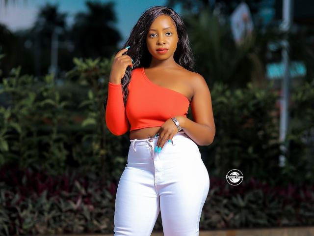 PrettyRebecca, con piel oscura, cabello negro largo, una blusa naranja con un solo hombro, jeans blancos de cintura alta, uñas azules y una pulsera de plata, de pie con confianza. Los senos son de tamaño medio, y las caderas son curvas..