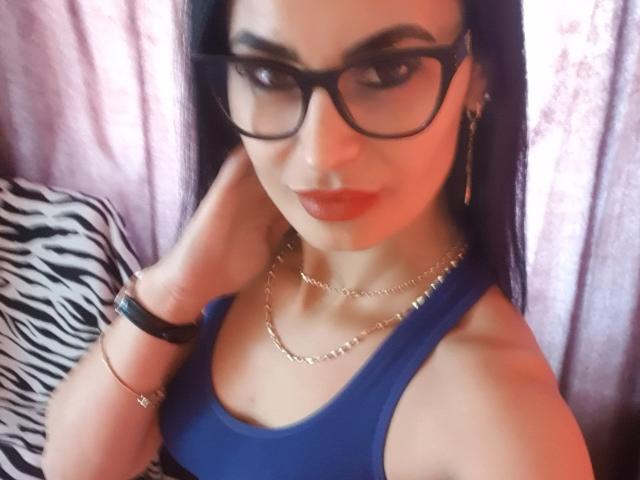 TheSunshine has dark hair, black glasses, red lipstick, and a blue top. She's wearing gold chains and black bracelets, with a zebra-patterned pillow in the background.