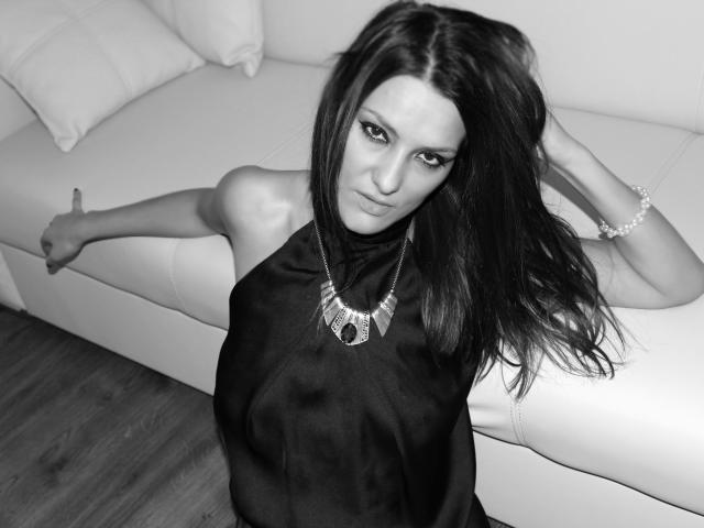 JoliDanielle with long dark hair, looking up, wearing a black halter dress, necklace, and bracelet, on a white couch, wooden floor, hand on head, one arm out.