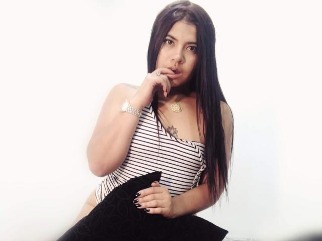 DulceCloe con lunghi capelli neri, maglietta a righe, che tiene un cuscino nero, con un dito vicino alla bocca, che appare sexy, orologio d'argento, collana d'oro, piccolo tatuaggio sul petto, sfondo bianco..