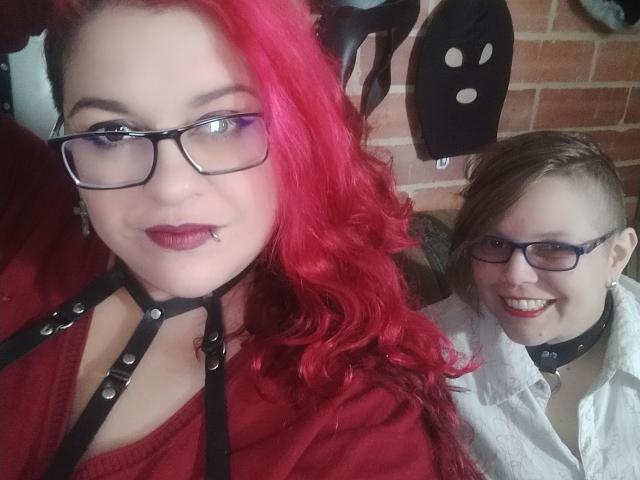 Deux jeunes femmes latino-américaines, aux cheveux roux et bruns, portant des lunettes et des colliers en cuir noir. Une Latina aux cheveux roux avec un piercing au nez, une autre Latina avec une chemise blanche, souriante. Les seins de la Latina aux cheveux roux sont visibles..