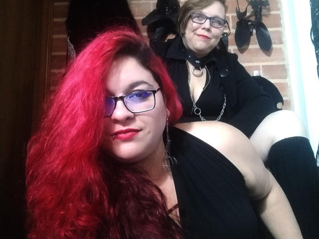 Ragazze latinoamericane con capelli rossi, occhiali e piercing al naso, che tengono un'altra ragazza latinoamericana con una maglietta nera, scollata e calze al ginocchio, entrambe vestite di nero, in una stanza poco illuminata..