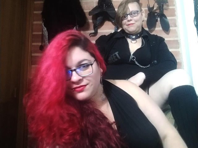 LatinoHardGirls with red hair and glasses, tits in a black top, sitting beside LatinoHardGirls with short blonde hair, glasses, black clothes, and a chain collar, showing a white dildo.