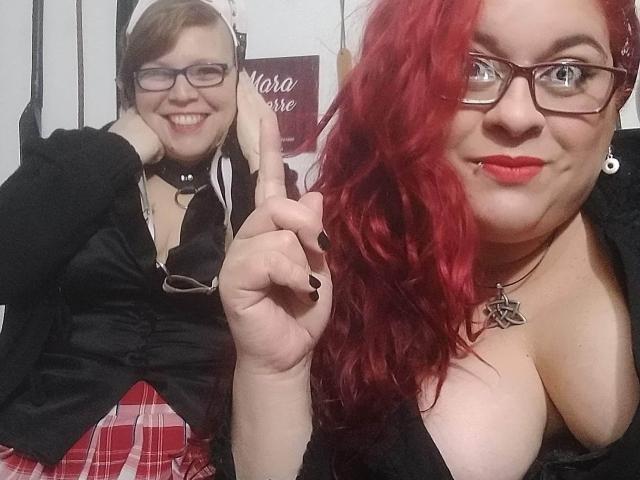 Two LatinoHardGirls with glasses, one with brown hair and the other with red, showing off cleavage in black tops and plaid skirts, making a peace sign. Red lipstick.