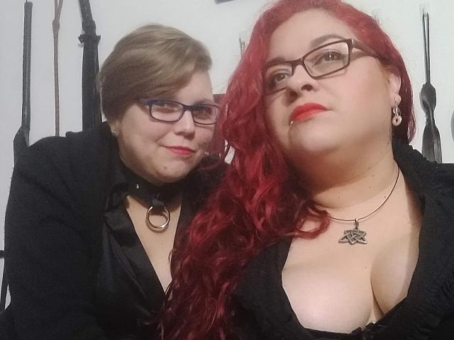 Deux jeunes femmes latino-américaines, aux cheveux roux et bruns, portant des lunettes, des tenues noires et avec un décolleté, se tiennent près l'une de l'autre, souriantes. La rousse a les cheveux bouclés, porte un collier avec un anneau et du rouge à lèvres rouge. Les seins sont visibles..