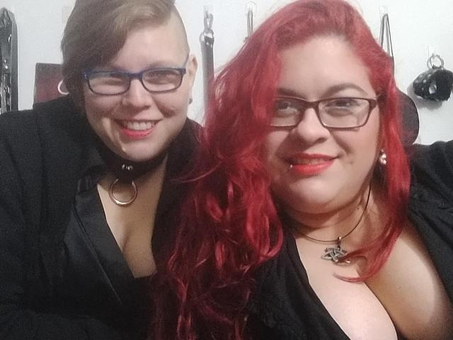 Deux jeunes femmes latino-américaines, portant des lunettes et du rouge à lèvres, dont l'une a les cheveux bruns et l'autre les cheveux roux, toutes deux vêtues de t-shirts noirs et de colliers ras du cou, montrant leurs seins..