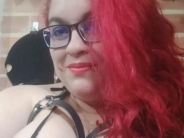 LatinoHardGirls mit roten Haaren, Brille und schwarzen Trägern, die ihre großen Brüste zur Schau stellen und in die Kamera lächeln. Sie hat einen niedlichen Nasenring und roten Lippenstift..