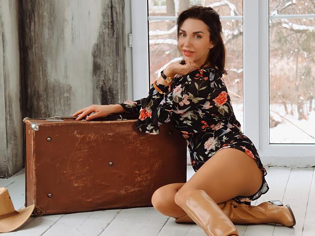 SimoniGrace con botas marrones, un vestido floral y medias color beige, arrodillada junto a una maleta de cuero marrón, mostrando su trasero, en una habitación con una ventana..