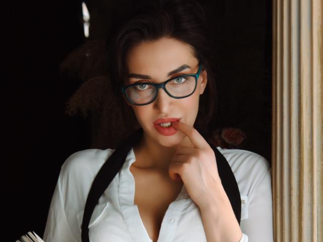SimoniGrace, met blauwe brillen, een witte blouse, zwarte bandjes en een vinger op haar rode lippen, heeft een schattige, sexy uitstraling, waarbij haar borsten licht zichtbaar zijn, ze heeft donkerbruin haar en een serieuze blik..